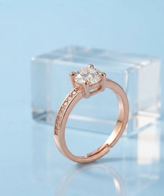 A rose gold engagement ring with a round-cut clear gemstone and a pavé band, displayed on a light blue reflective surface with a clear acrylic block in the background.