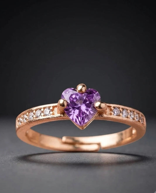 A rose gold adjustable ring featuring a heart-shaped purple amethyst gemstone in the center, with small clear stones set along the band, shown on a dark gray background.