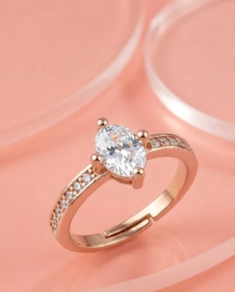 A rose gold engagement ring with a pear-cut clear center stone and small pavé-set stones along the band, displayed on a soft peach-pink background.
