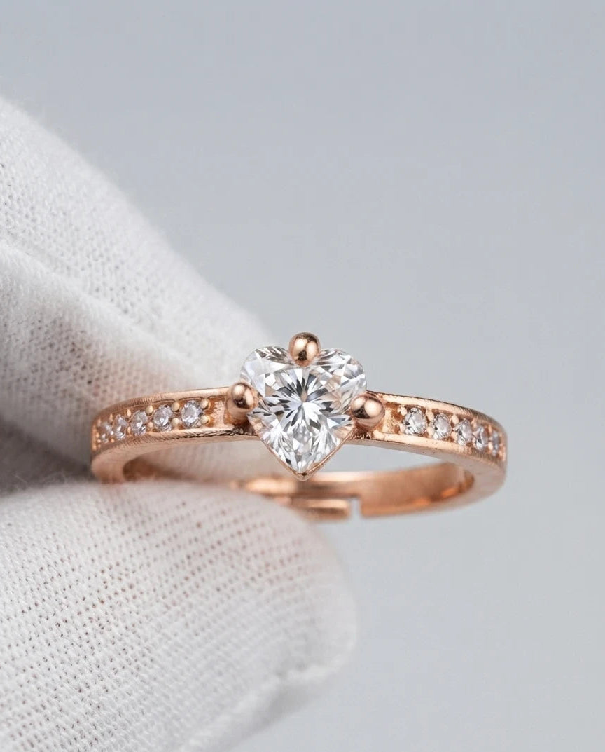A rose gold ring with a heart-shaped diamond center stone and small pavé diamonds on the band, resting on a white textured fabric surface.