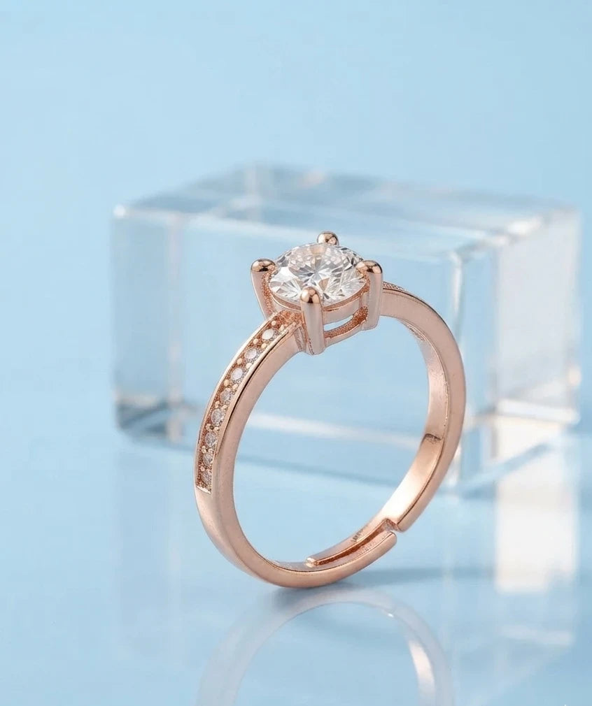 A rose gold engagement ring with a round-cut clear gemstone and a pavé band, displayed on a light blue reflective surface with a clear acrylic block in the background.