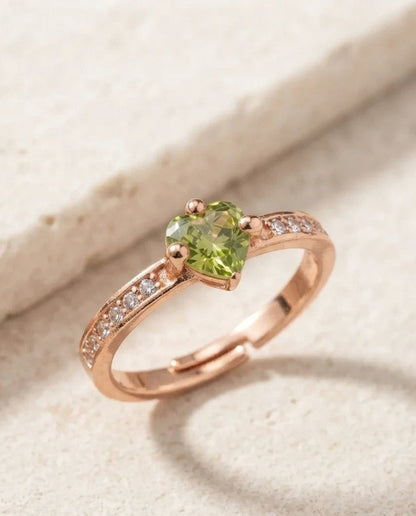 A rose gold ring featuring a heart-shaped green peridot gemstone and small clear accent stones along the band, displayed on a textured beige stone surface.