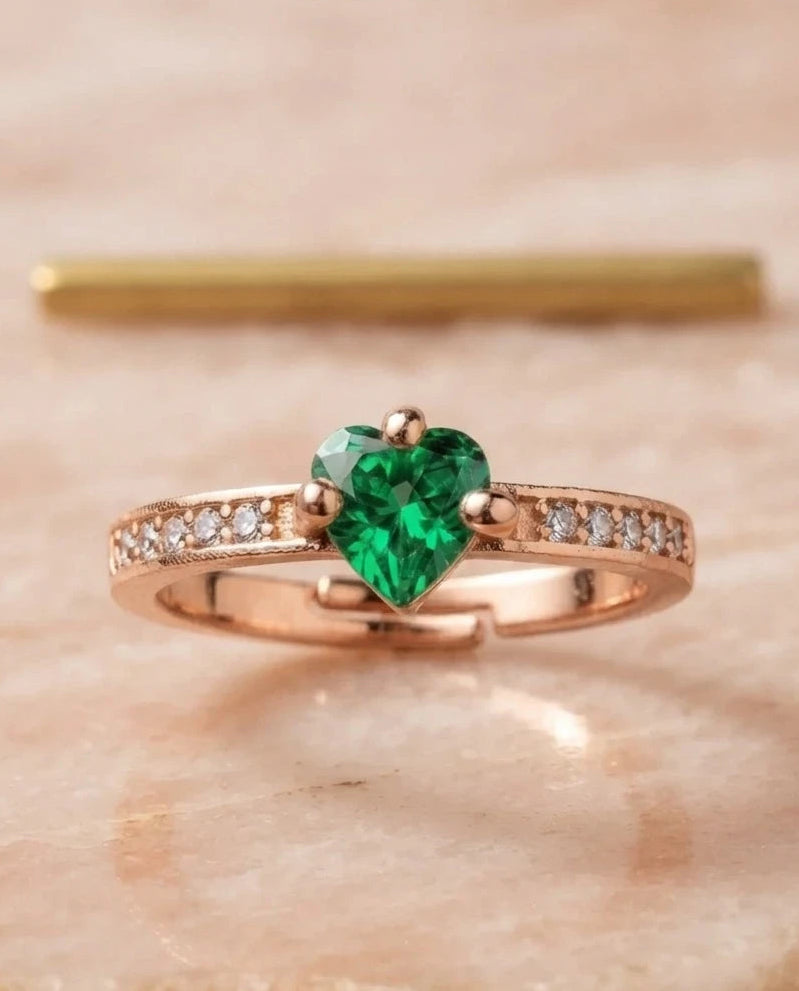 A rose gold ring featuring a heart-shaped green emerald center stone and pavé-set clear accent stones on the band, displayed on a light beige stone surface.
