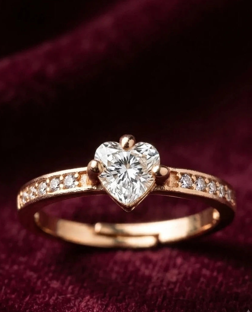 A rose gold ring featuring a heart-shaped clear gemstone in the center, with small pavé-set diamonds along the band, displayed on a deep burgundy velvet background.