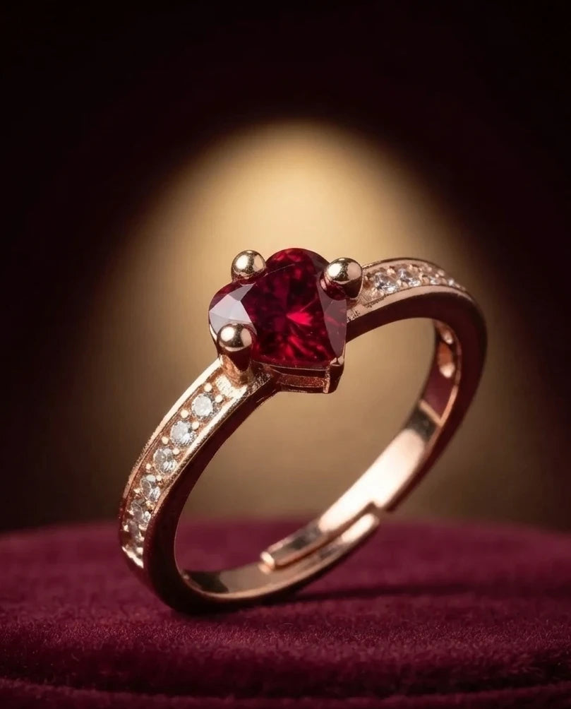 A rose gold ring with a heart-shaped red gemstone and small clear accent stones on the band, resting on a dark burgundy velvet surface with a warm spotlight in the background.