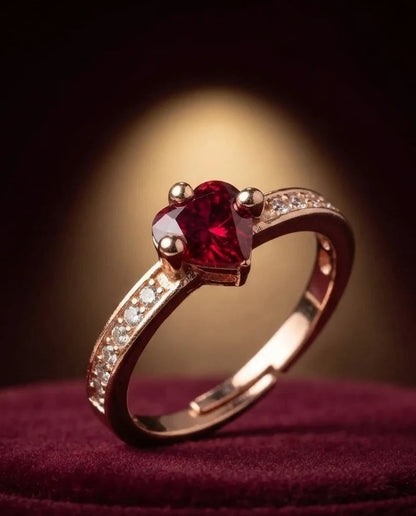 A rose gold ring with a heart-shaped red gemstone and small clear accent stones on the band, resting on a dark burgundy velvet surface with a warm spotlight in the background.