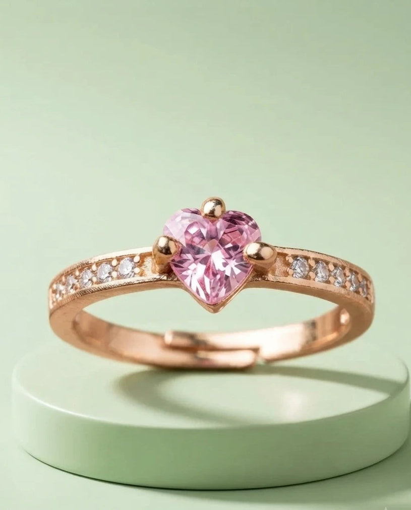A rose gold ring with a heart-shaped pink gemstone and small clear accent stones on the band, displayed on a light green circular pedestal against a matching light green background.